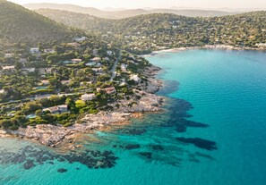 Une vue aérienne d’une ville côtière, avec des maisons le long du littoral, des eaux turquoise et limpides, et des collines environnantes.