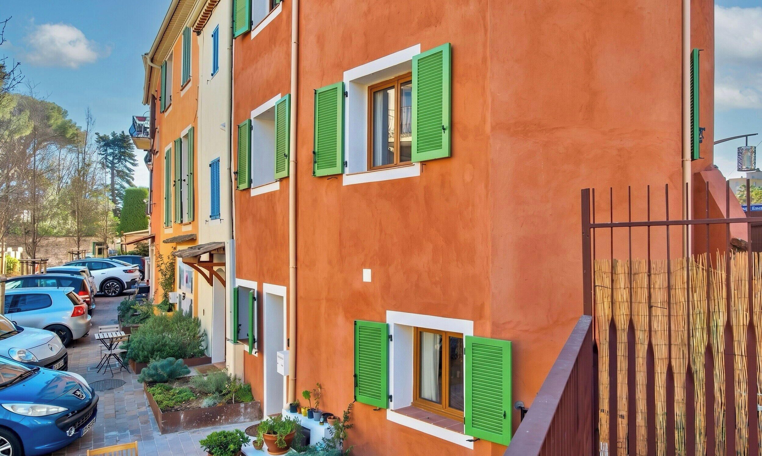 A multi-story building with green shutters, a parking area with cars, and a bamboo fence.