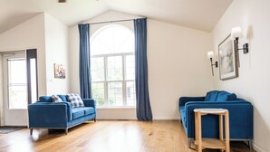 Een ruime woonkamer met twee blauwe banken, een houten vloer, een groot raam met gordijnen en een kleine tafel.
