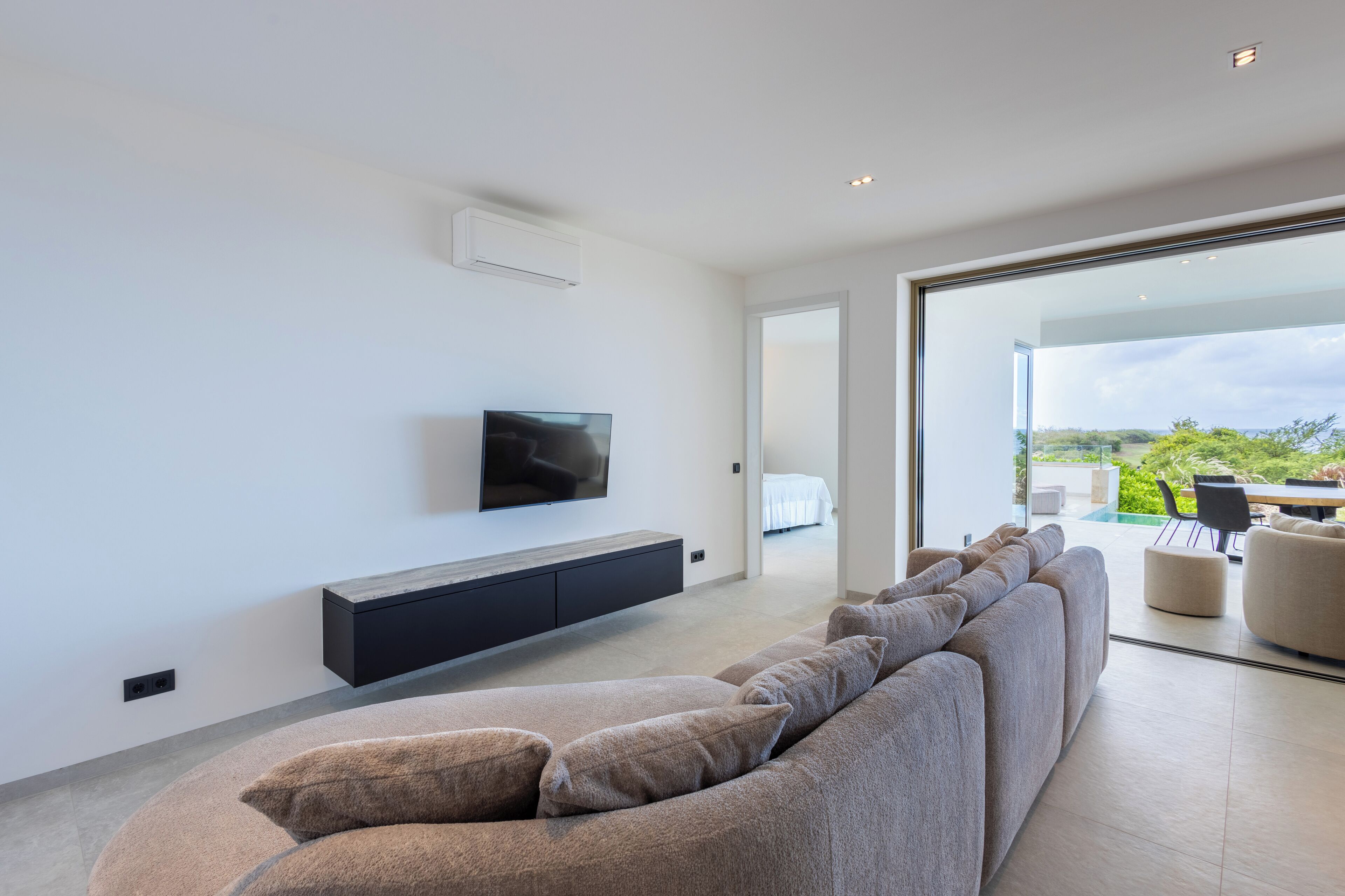 Una sala de estar moderna con un sofá grande, un televisor de pantalla plana montado en la pared y un ventanal corredizo que da a un área exterior.