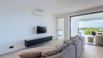 A modern living room with a large sofa, a flat-screen TV mounted on the wall, and a sliding glass door leading to an outdoor area.