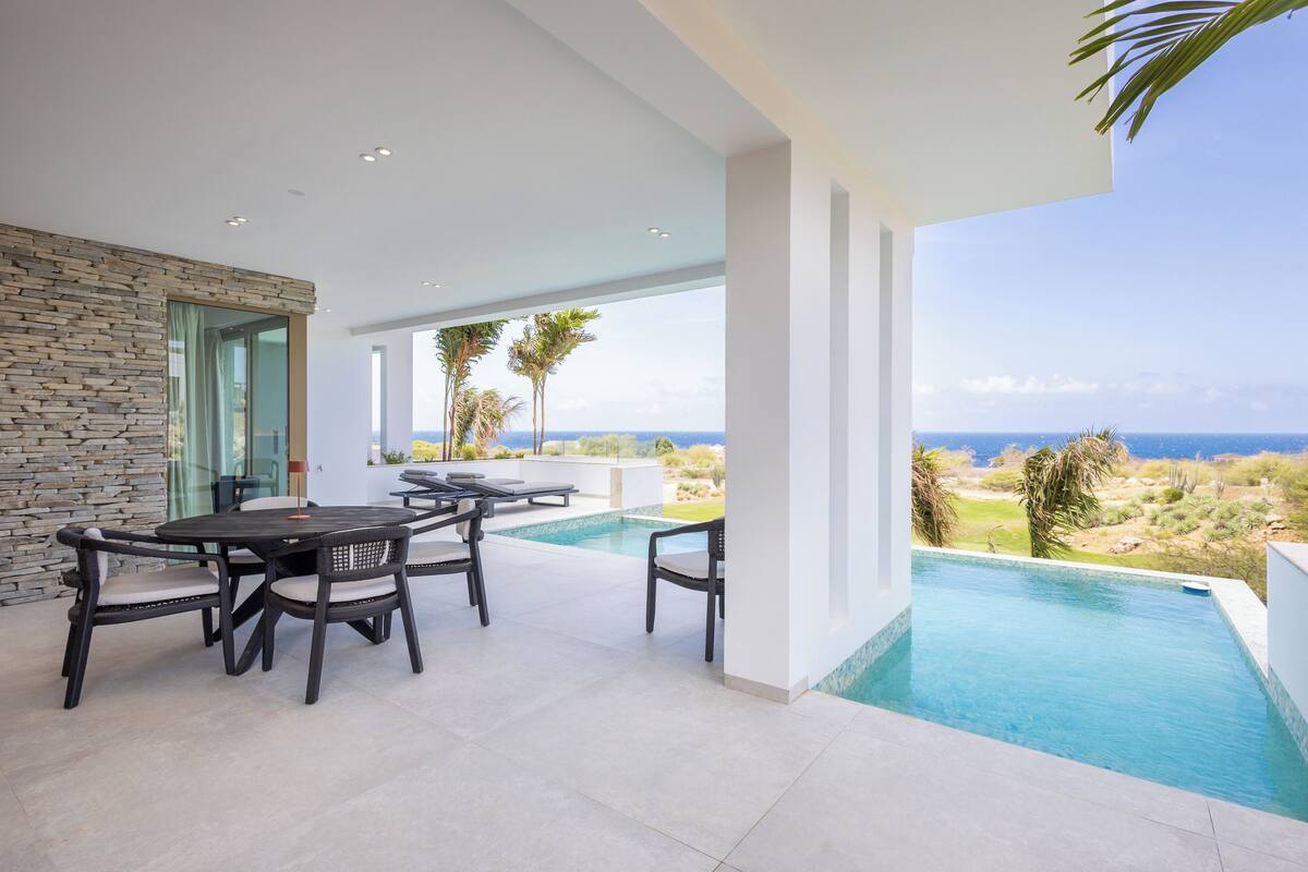 A modern outdoor pool area with a stone wall, a dining set, and a view of the ocean.