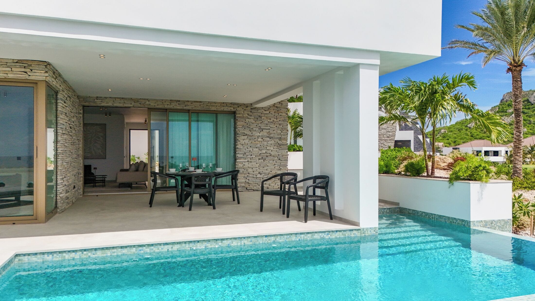 A modern outdoor pool area with a dining setup, stone walls, and a view of a residential area.