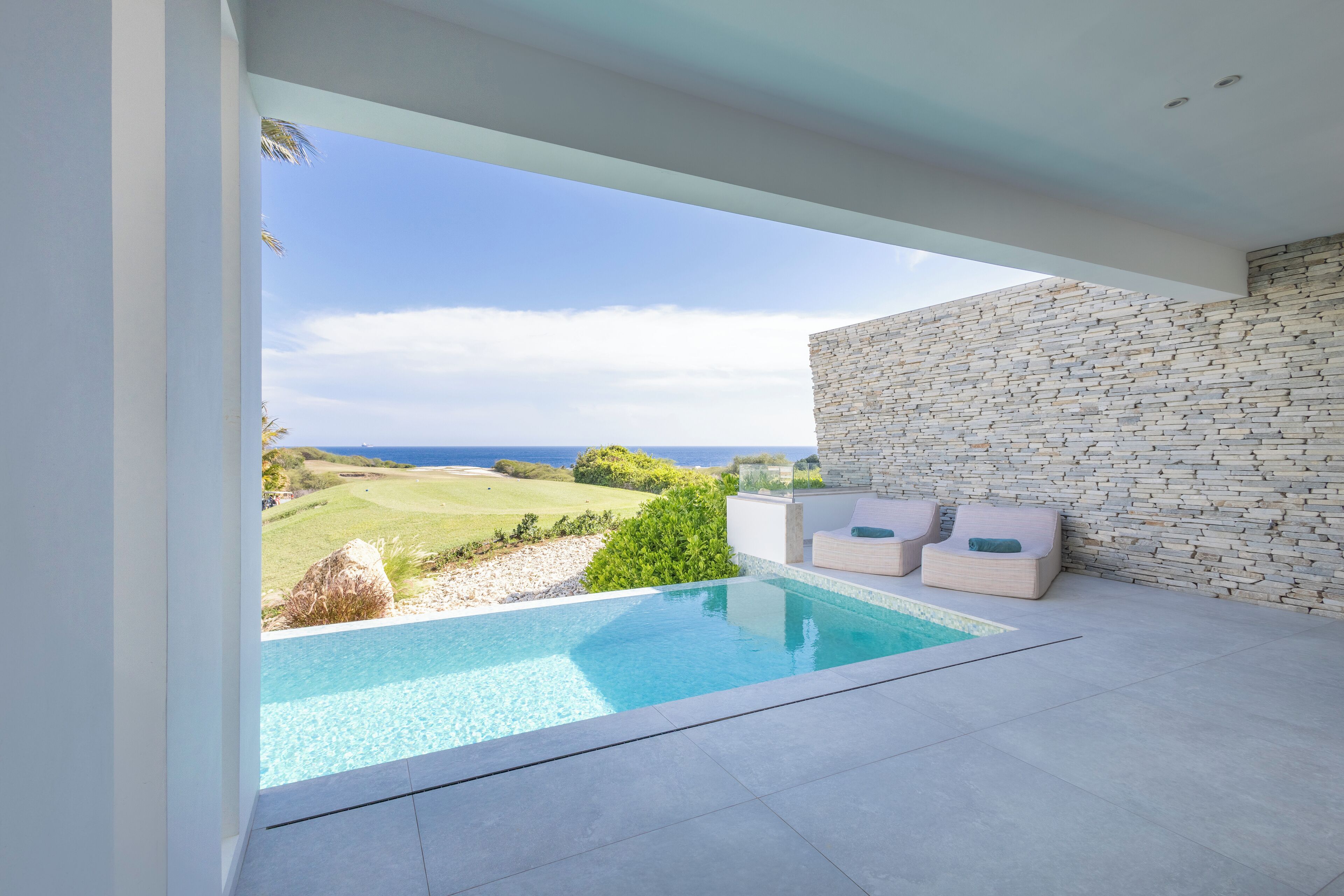 A modern outdoor pool area with a stone wall and two lounge chairs, overlooking a coastal landscape.