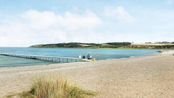 A beach with a pier extending into calm waters, a grassy area in the foreground, and a hilly landscape in the background.