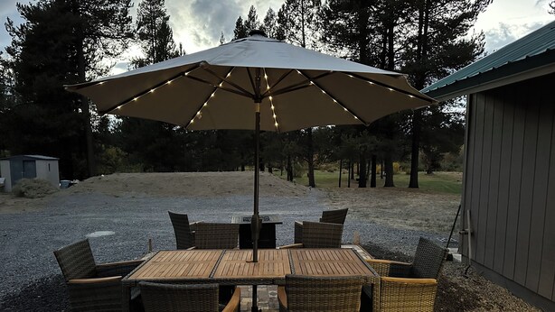 An outdoor patio with a table and chairs, an umbrella, and a gravel area with trees in the background. - Charming 2-bedroom cottage waterfront on the little Deschutes River  La Pine, OR (La Pine)