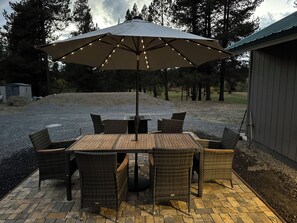An outdoor patio with a table and chairs, an umbrella, and a gravel area with trees in the background. - Charming 2-bedroom cottage waterfront on the little Deschutes River  La Pine, OR (La Pine)