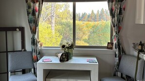 A room with a white desk and two grey chairs, a window with curtains, and a view of trees outside. - Charming 2-bedroom cottage waterfront on the little Deschutes River  La Pine, OR (La Pine)