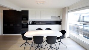 A modern kitchen with white cabinets, a black countertop, and a dining area with a white table and black chairs. - Apartment in Oostende Near the Beach (De Haan)