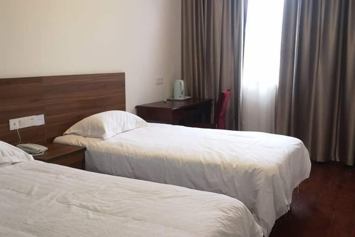 A hotel room with two beds, a wooden headboard, a bedside table with a phone, a lamp, and a view of the outside through curtains.