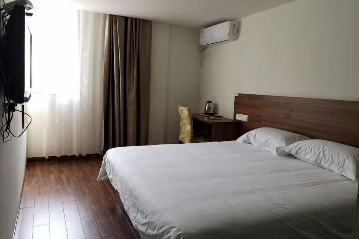A hotel room with a wooden floor, a bed with white linens, a bedside table with a lamp, and a window with curtains.