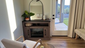 Un salon moderne avec un plancher en bois, un canapé, une petite table ronde avec une bougie, une cheminée, un grand miroir et une vue sur un jardin, visible à travers une porte vitrée.