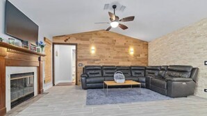 Een moderne woonkamer met een open haard, een zwarte leren zithoekbank, een houten accentmuur en een plafondventilator.