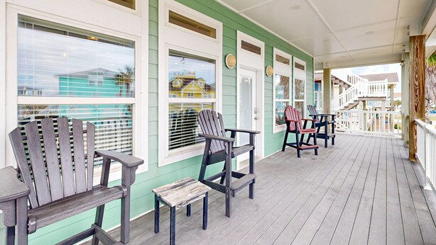A covered porch with wooden chairs and a small table, a white railing, and a view of houses in the background. - Pet Friendly, Golf Cart Included, Walk to the Beach (Port Aransas)
