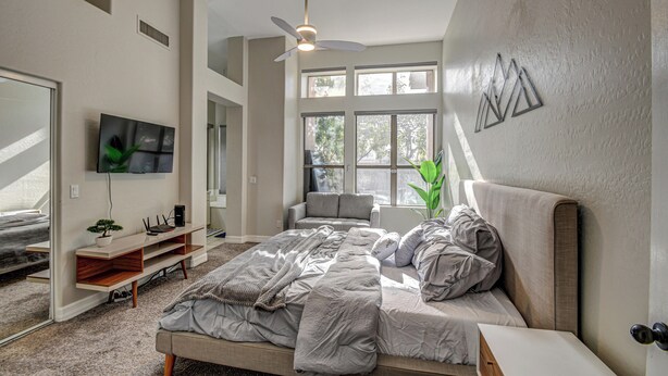 Un dormitorio con cama, televisión, ventilador de techo y ventanas amplias.
