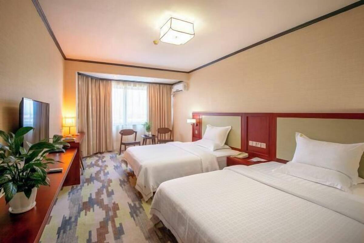A hotel room with two beds, a desk with a TV, a chair, a lamp, and a potted plant.