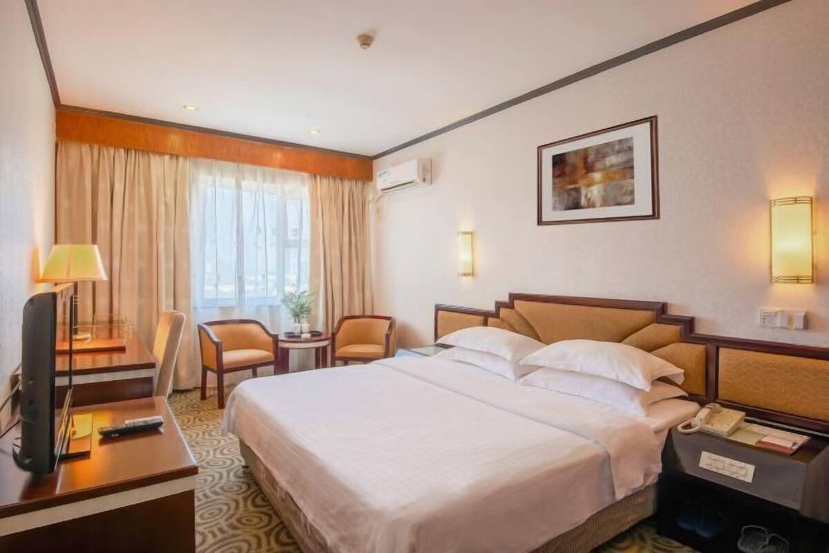A hotel room with a bed, a desk with a lamp, a chair, a bedside table with a phone, and a wall-mounted air conditioner.