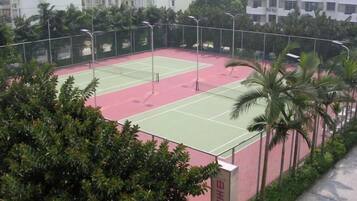 An outdoor tennis court surrounded by greenery and a building in the background.