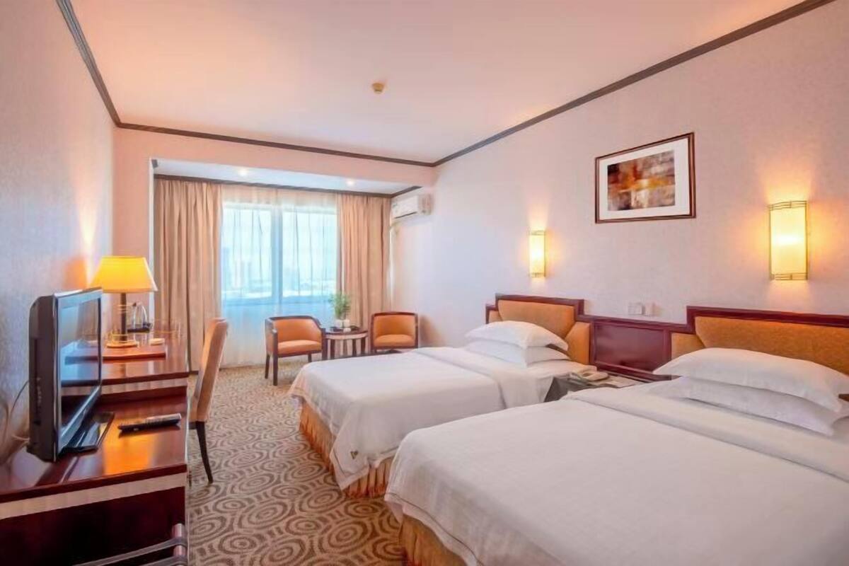 A hotel room with two beds, a desk with a TV, a chair, a small table, and a window with curtains.