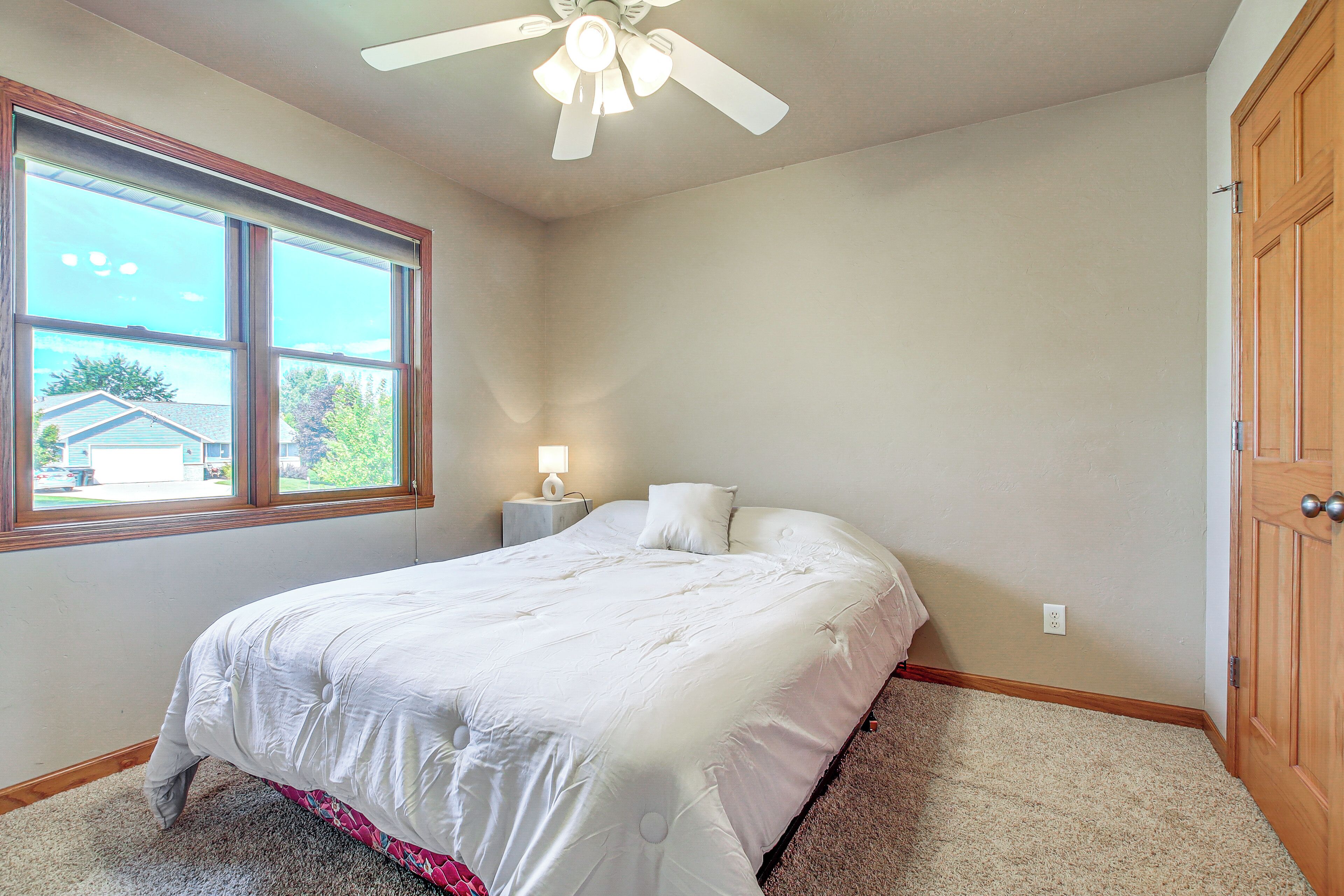 A bedroom with a bed, a ceiling fan, a nightstand with a lamp, a window, and a door.