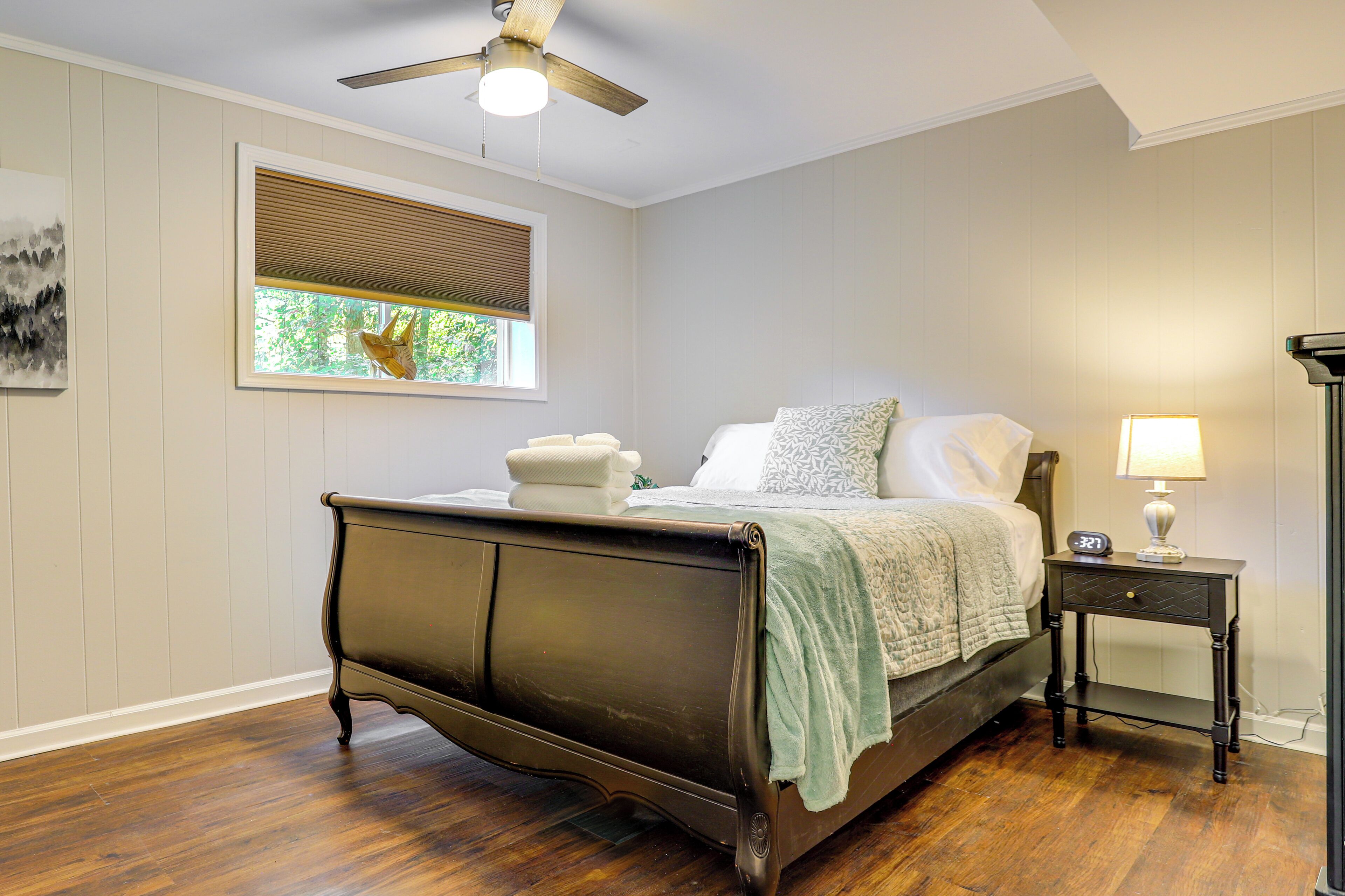 A bedroom with a dark wooden bed, a bedside table with a lamp, a window with blinds, and a ceiling fan.