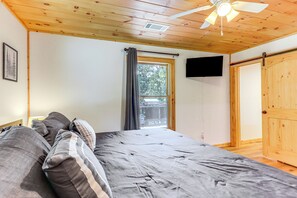 Une chambre à coucher avec un lit, un ventilateur de plafond, une fenêtre avec des rideaux, un téléviseur fixé au mur et une porte en bois.