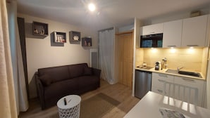 Ett kompakt bostadsutrymme med en soffa, ett litet runt bord, en köksdel med mikrovågsugn, diskho och skåp, samt en dörr med en gardin.