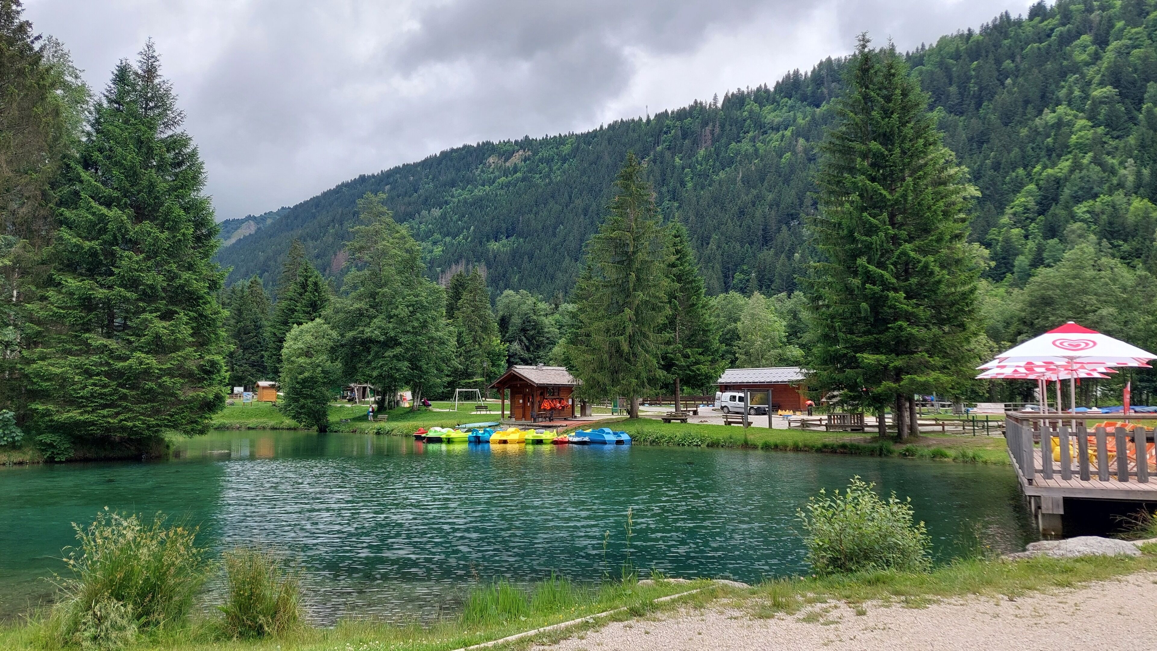 A lakeside view with a wooden dock, paddle boats, and cabins in a forested area.