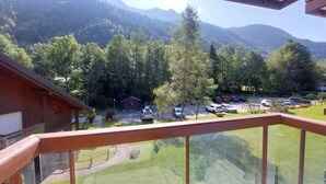 Vista desde un balcón sobre un estacionamiento con varios coches, un edificio pequeño y montañas cubiertas de bosque al fondo.