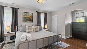 A bedroom with a metal bed frame, a bedside table with a lamp, a dresser, and a large window with curtains. - Downtown - Sleeps 8 - We Love Doggies! (Wilmington)