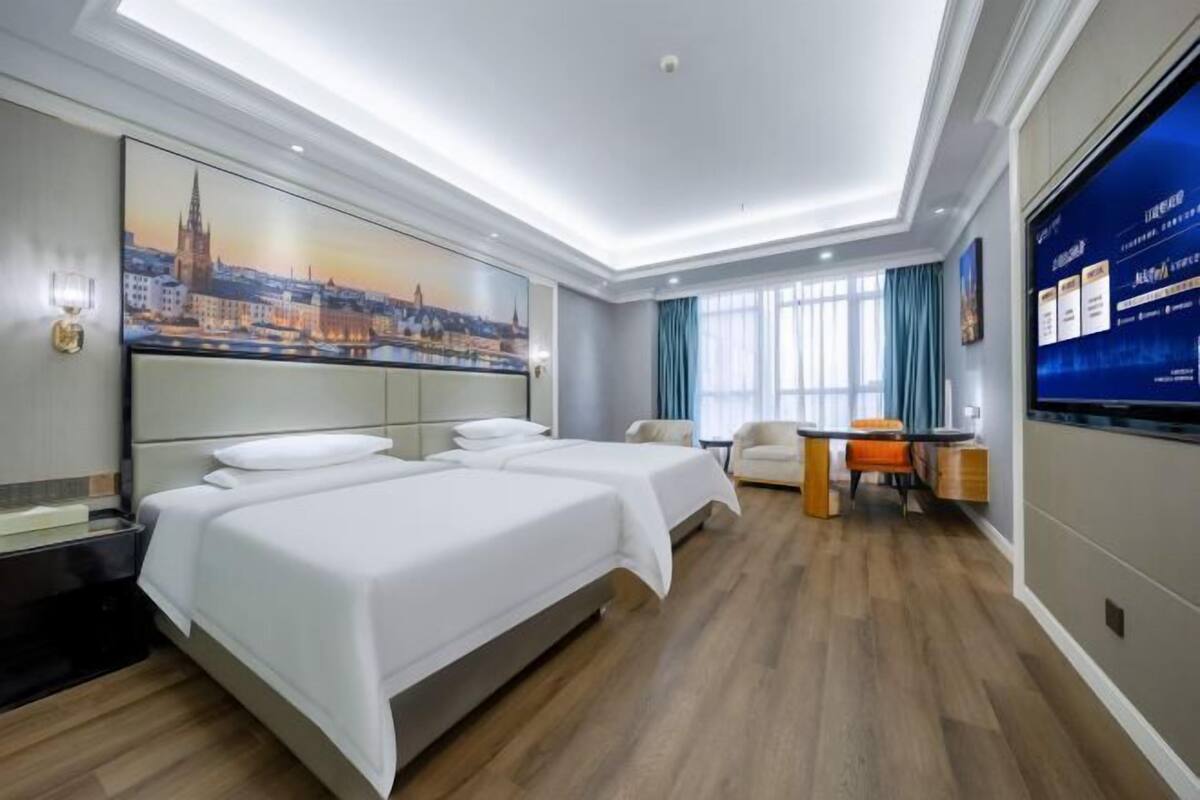 Une chambre d’hôtel avec un grand lit, un bureau, une chaise, une télévision et un tableau représentant un paysage urbain accroché au mur.