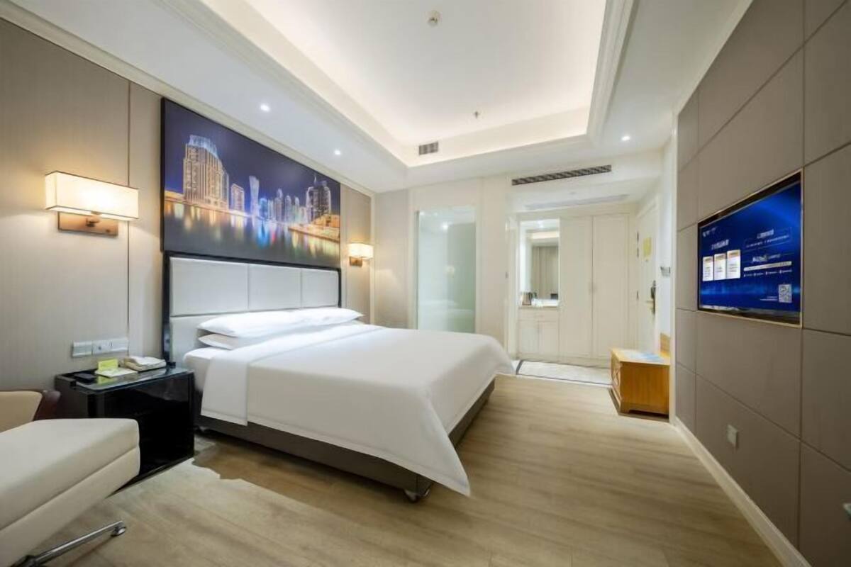 Une chambre d’hôtel moderne, dotée d’un grand lit, d’un bureau, d’une chaise, d’une télévision et d’un tableau représentant un paysage urbain accroché au mur.