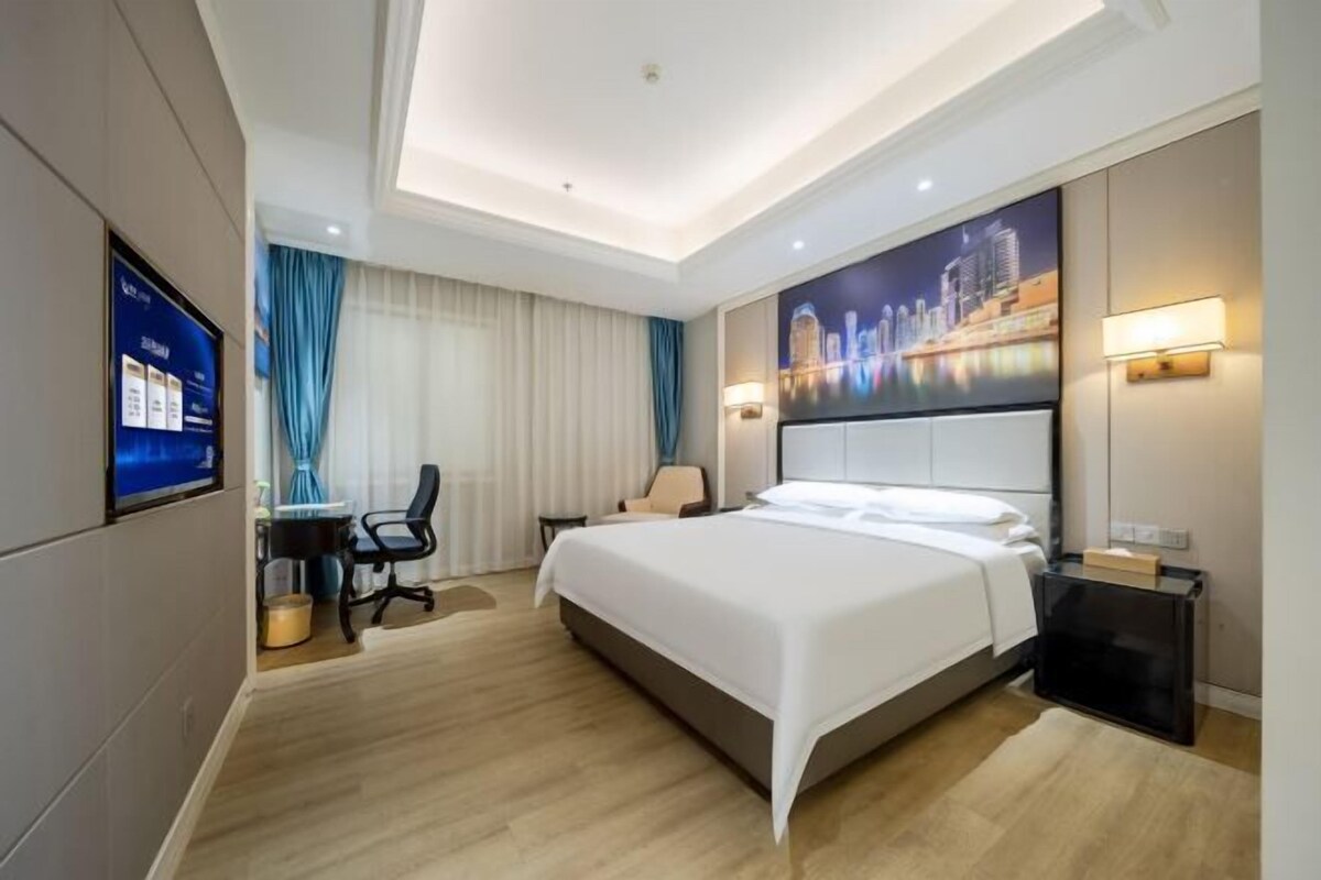 Une chambre d’hôtel moderne, dotée d’un grand lit, d’un bureau avec une chaise, d’une télévision et d’un tableau représentant un paysage urbain accroché au mur.