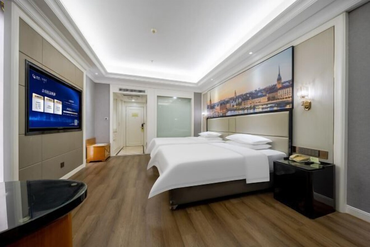 Une chambre d’hôtel avec un lit, un bureau, une télévision et un tableau représentant un paysage urbain.