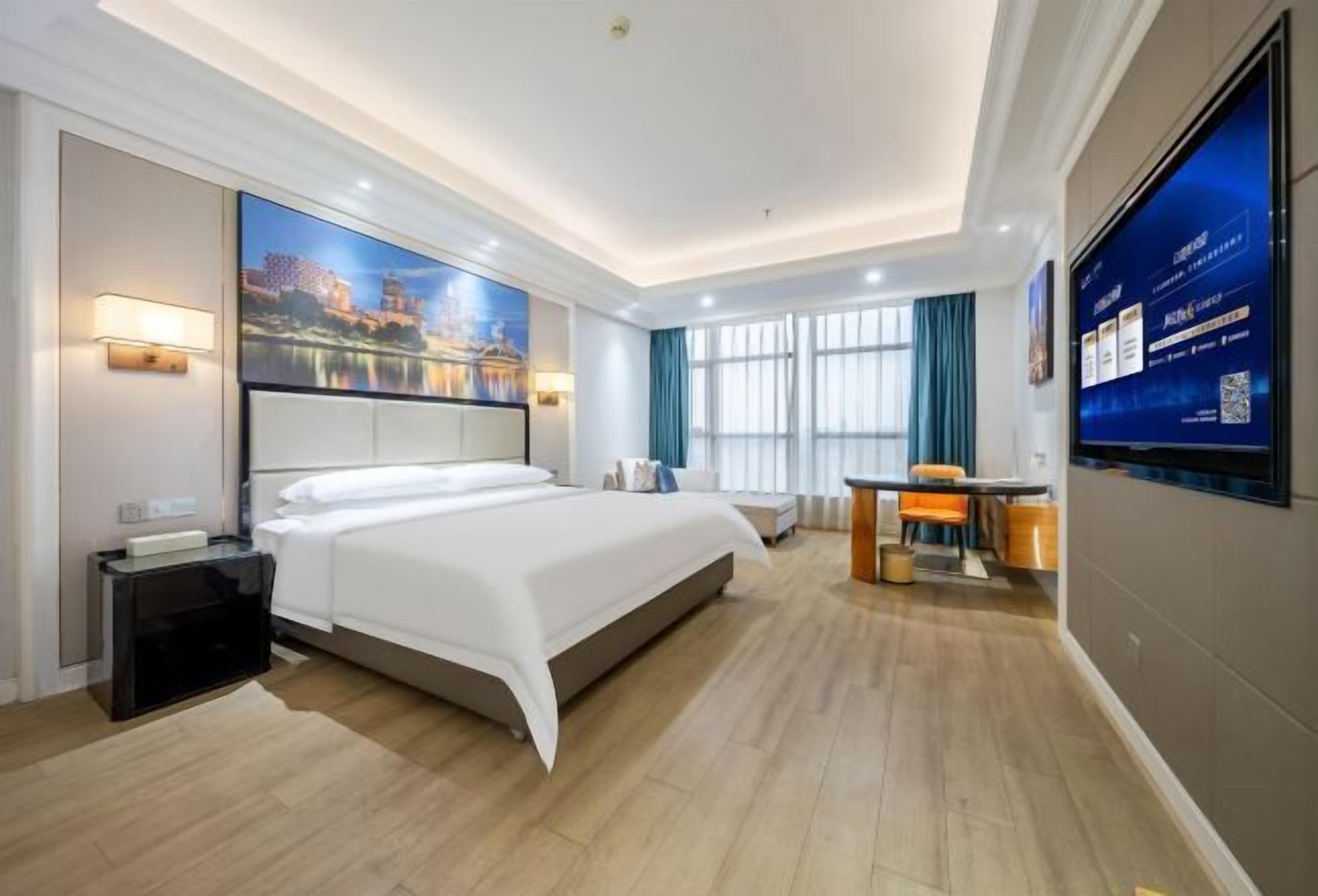 A hotel room with a large bed, a desk, a flat-screen TV, and a cityscape painting on the wall.