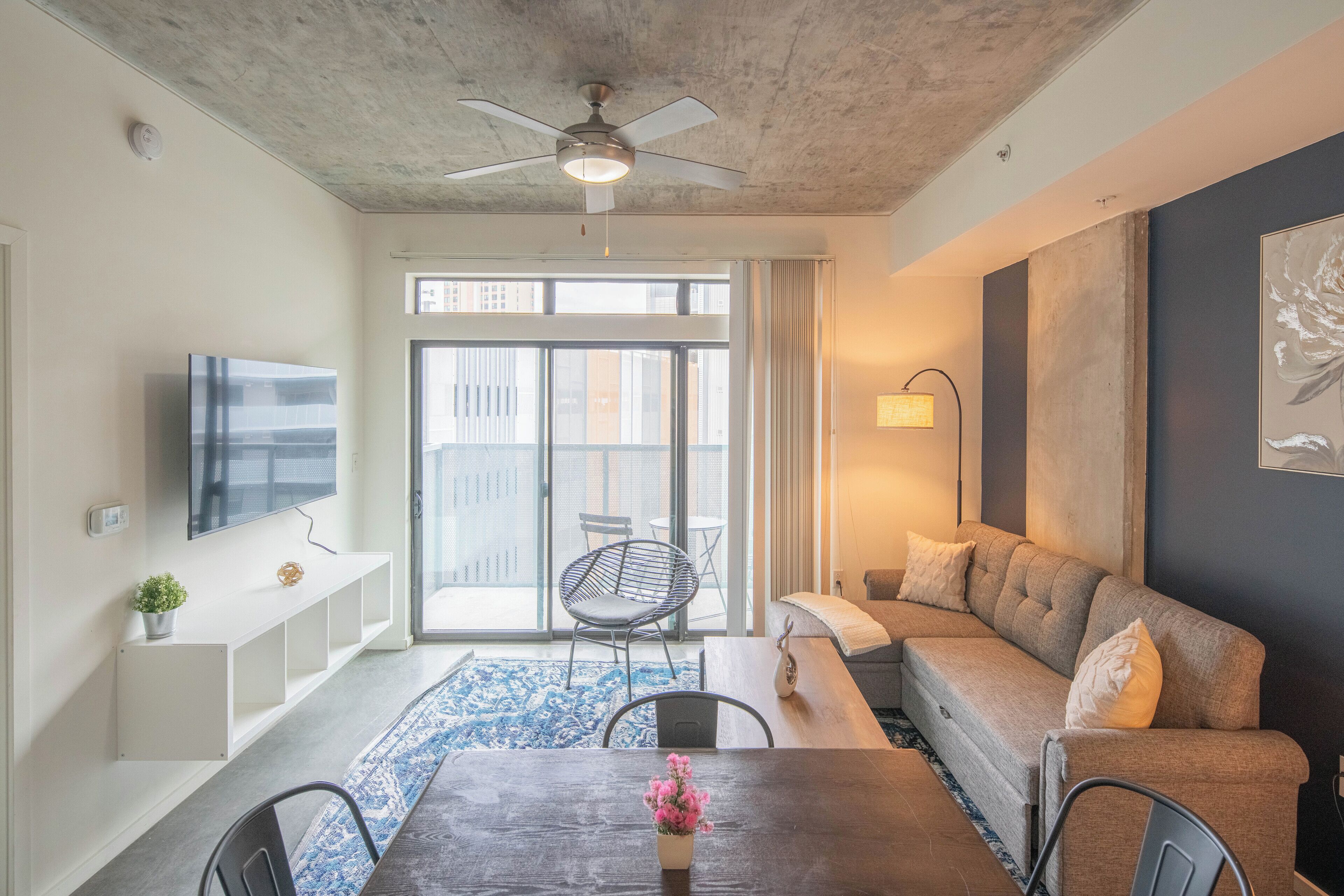 A modern living room with a ceiling fan, a flat-screen TV, a sofa, a coffee table with a vase of flowers, and a large window with a view of buildings.