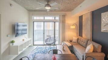 A modern living room with a ceiling fan, a flat-screen TV, a sofa, a coffee table with a vase of flowers, and a large window with a view of buildings.