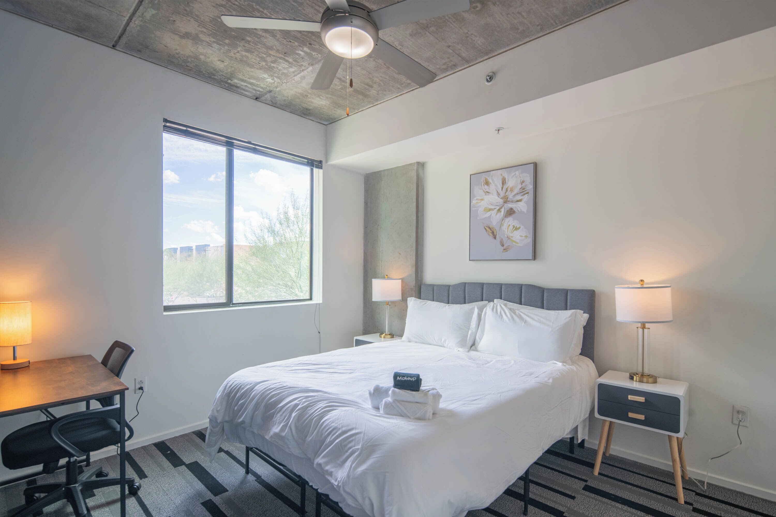 Una habitación de hotel moderna con una cama grande, dos mesitas de noche con lámparas, un escritorio con silla y una ventana con vistas a zonas verdes.