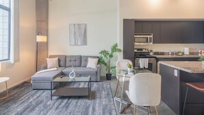 Ein modernes Wohnzimmer mit einem grauen Ecksofa, einem Glastisch und einem kleinen Beistelltisch. Eine Topfpflanze und ein Gemälde sind an der Wand zu sehen.