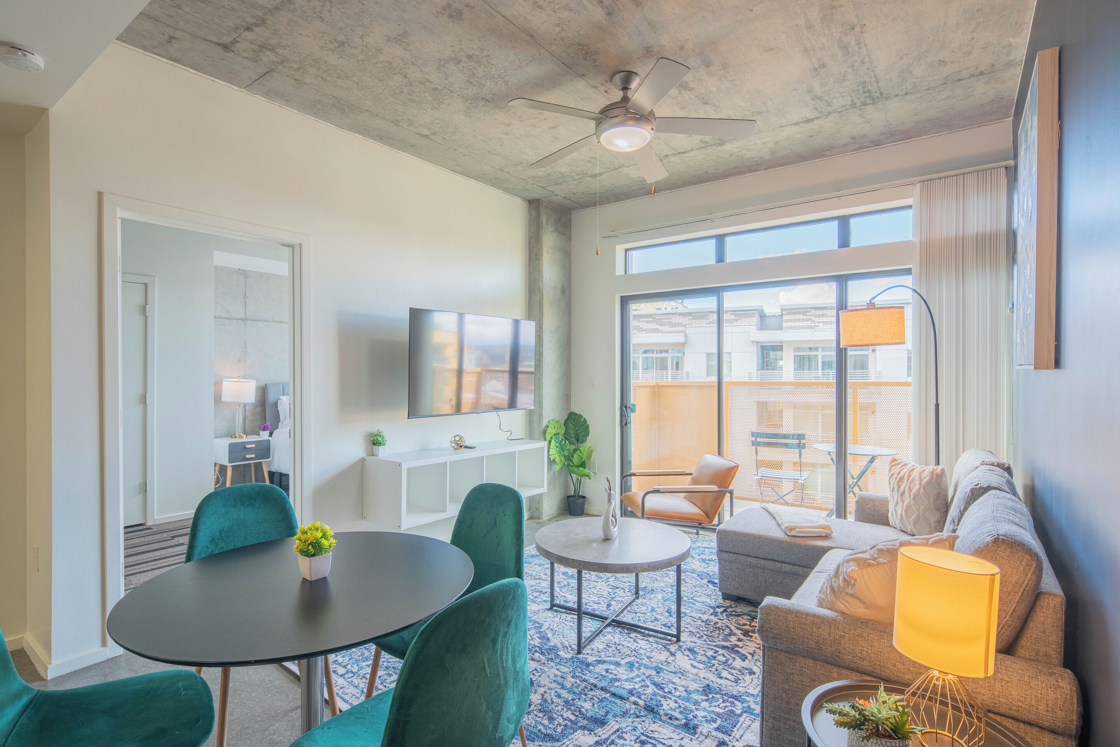 Una sala de estar moderna con un sofá, una mesa de comedor con sillas, un televisor y un amplio ventanal con vistas a edificios.