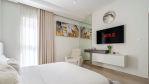 Et moderne hotelværelse med en stor seng, et siddeområde med en stol, et vægmonteret fjernsyn og kunst på væggen.