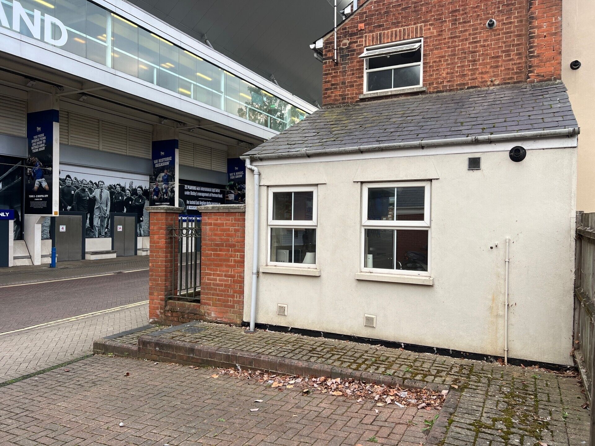 A small white building with two windows and a brick roof, adjacent to a larger building with a glass facade.