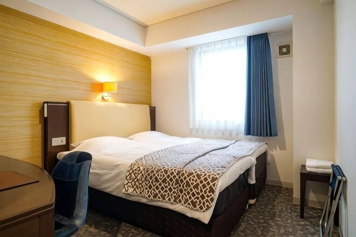 A hotel room with a bed, a desk, a lamp, a window with curtains, and a small table.