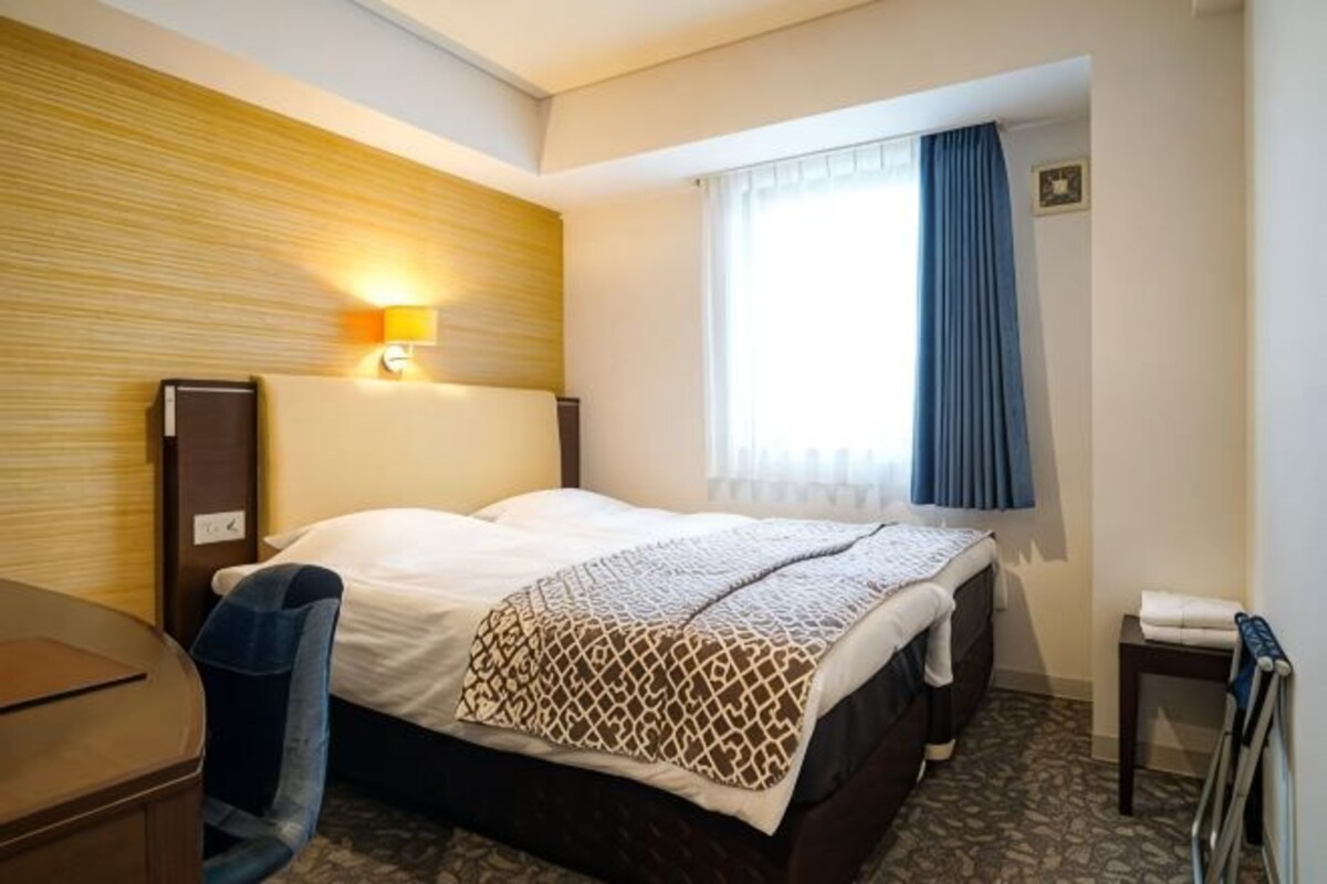 Ett hotellrum med en säng, ett skrivbord, en lampa, ett fönster med gardiner och ett litet bord.