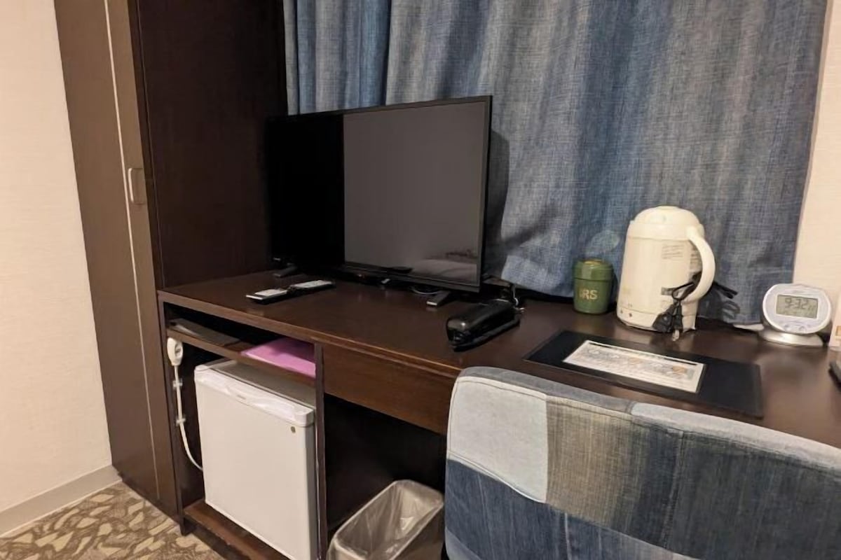 Ett hotellrum med ett skrivbord, en tv, en kaffebryggare, en papperskorg och ett litet kylskåp.