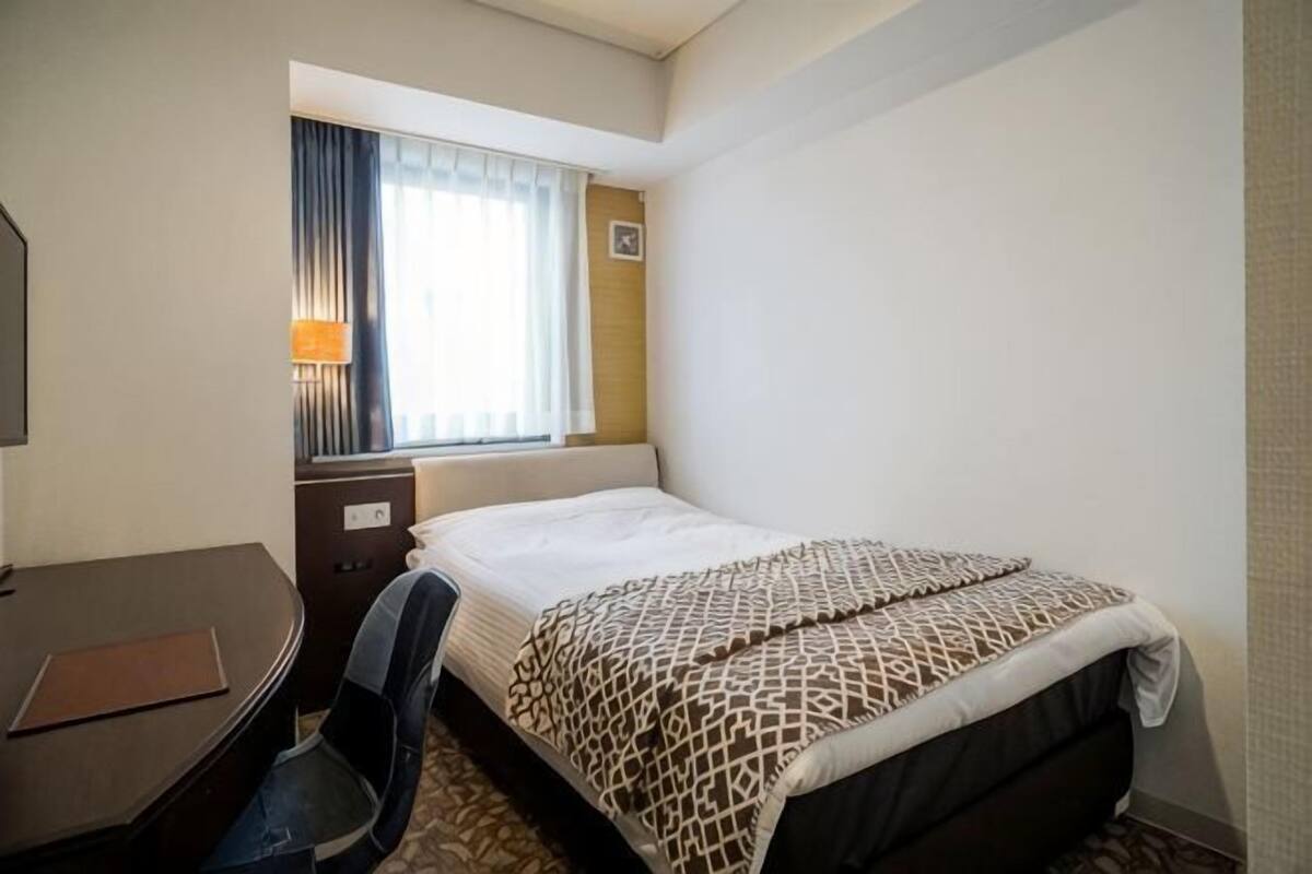 A hotel room with a bed, desk, chair, lamp, and window with curtains.