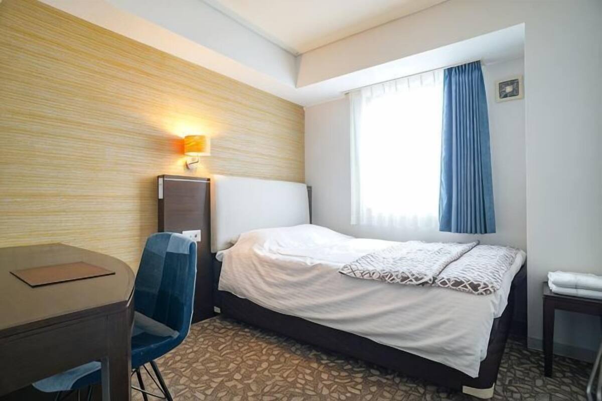 A hotel room with a bed, desk, chair, lamp, and window with curtains.