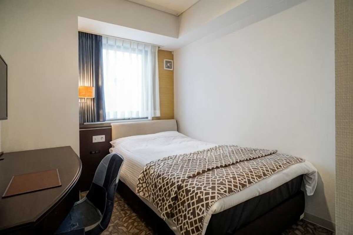 Hotellihuone, jossa on sänky, työpöytä, tuoli, lamppu ja ikkuna, jossa on verhot.