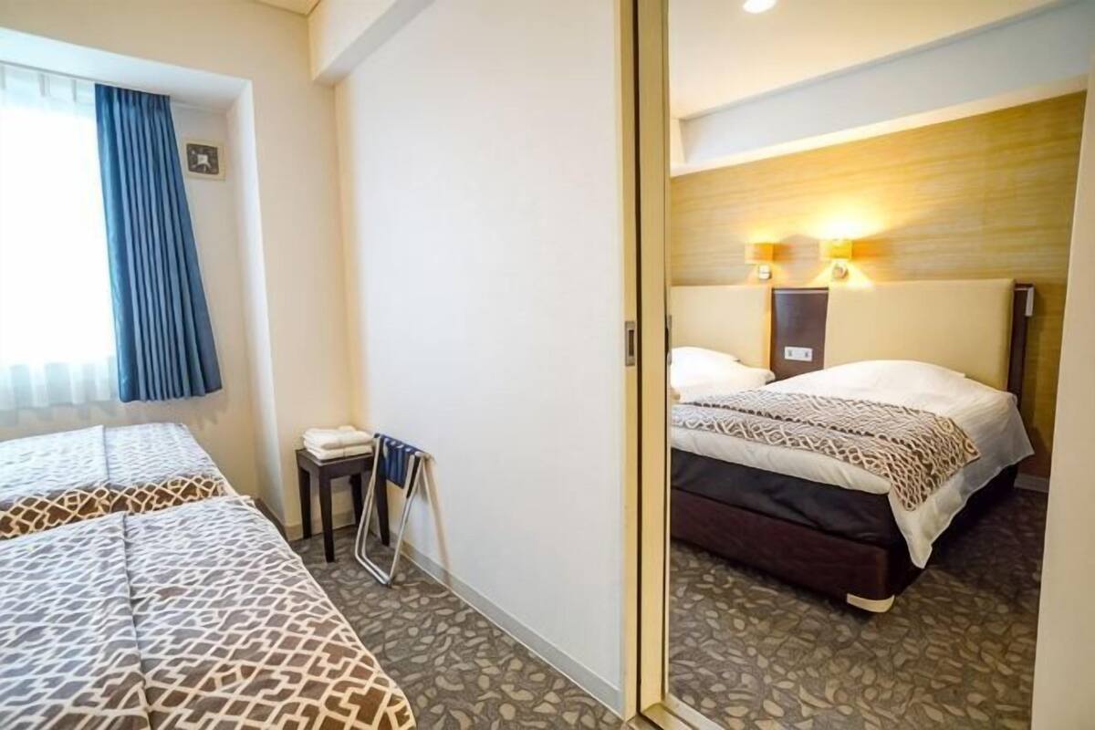 A hotel room with two beds, a small table, a chair, and a window with curtains.