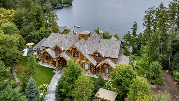 A large log cabin with a stone foundation, surrounded by lush greenery and a lake view.